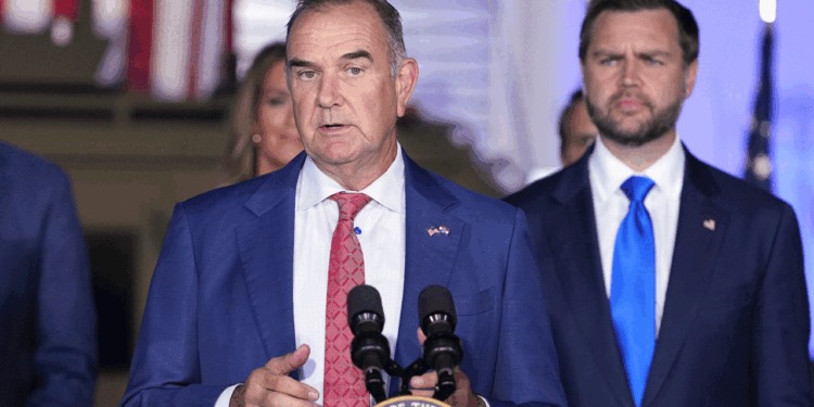 A man in a blue suit speaks at a podium with microphones, while another man in a suit stands behind him. An American flag is visible in the background, suggesting this event may be taking place in Missouri.