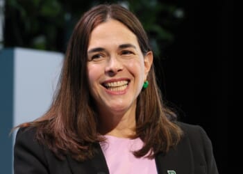 A woman with long brown hair, wearing a black blazer and pink top, smiles while seated indoors. Green plants are visible in the background.