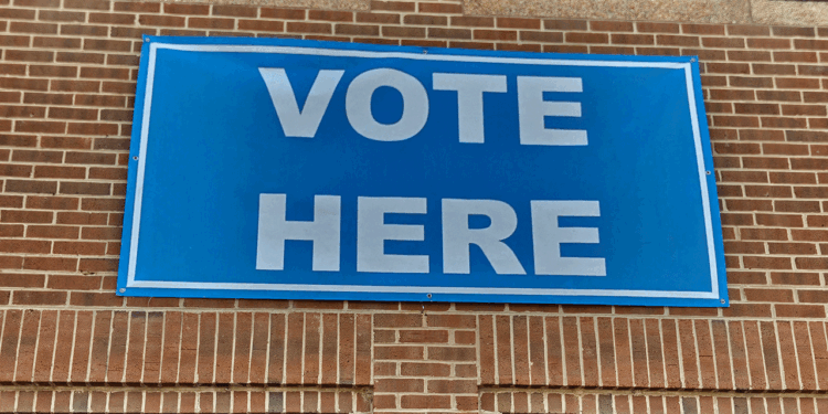 A blue sign with white letters reads VOTE HERE and is attached to a brick wall, indicating a polling location.