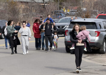 A group of adults and children walk together on a street near parked cars. One woman in front carries a child. Trees and emergency vehicles are in the background.