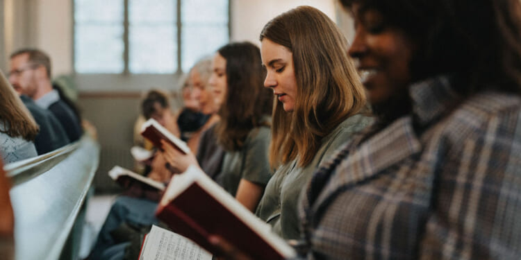 People sit in church pews reading hymnals or songbooks. The focus is on a young woman with brown hair, while others are also engaged in reading. The scene is well-lit, and a window is visible in the background.