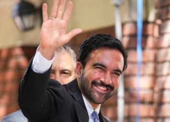 A man in a suit smiles and waves, standing outdoors near a brick wall. Another person is partially visible behind him.