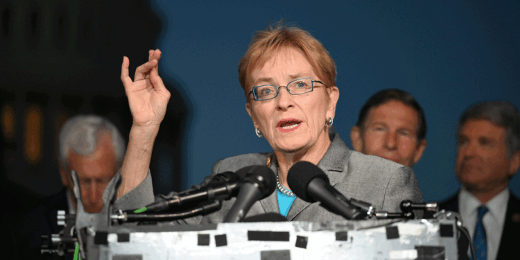 Marcy Kaptur in a gray suit and glasses speaks at a podium with microphones, gesturing with her hand. Several men in suits stand behind her. The background is dark, with a blurred building visible on the left.