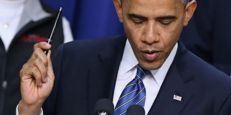 Barack Obama in a suit and striped blue tie, reminiscent of President Obama holds a pen up while speaking at a podium with microphones. An American flag pin is visible on the suit jacket.