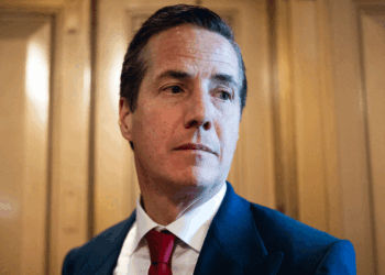 A man in a dark suit, white shirt, and red tie stands in front of wooden panels, looking slightly to his right with a neutral expression.