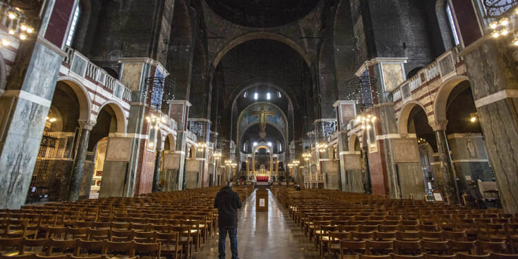 A person stands alone in the aisle of a grand, dimly lit cathedral with rows of empty wooden chairs and a large cross at the altar ahead.