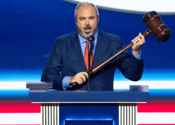 A man in a blue suit and red tie stands at a podium, holding a large wooden gavel and speaking into a microphone, with a blue and red background behind him.