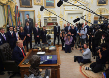 Donald Trump sits at the Resolute Desk in the Oval Office, surrounded by officials and media, with microphones and cameras pointed toward him. Portraits and gold decor line the walls.