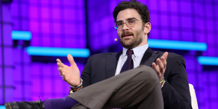 A man in a suit and tie sits on stage with a microphone headset, gesturing with his hands while speaking. The background is purple with blue horizontal accents.