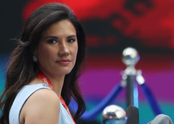 A woman with long dark hair wearing a light blue dress and a red lanyard looks toward the camera, with a blurred background of blue and red hues.