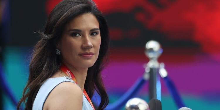 A woman with long dark hair wearing a light blue dress and a red lanyard looks toward the camera, with a blurred background of blue and red hues.