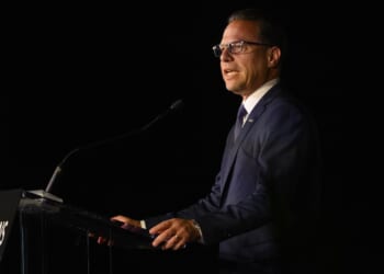 A man in a navy suit and glasses speaks at a podium with a microphone against a dark background. The podium sign reads INNOVATIONS PITTSBURGH..