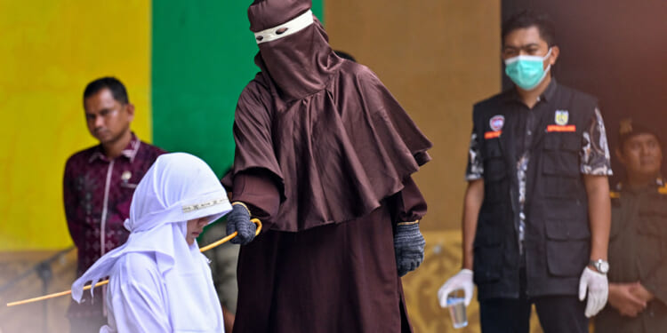 A person in brown robes prepares to cane a kneeling individual in white while uniformed officials and onlookers stand nearby, one wearing a face mask.