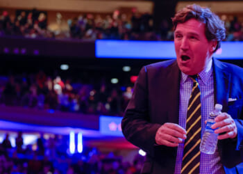 Tucker Carlson in a suit and striped tie, holds a water bottle as he stands in a crowded auditorium with colorful lights and a blurred audience in the background.