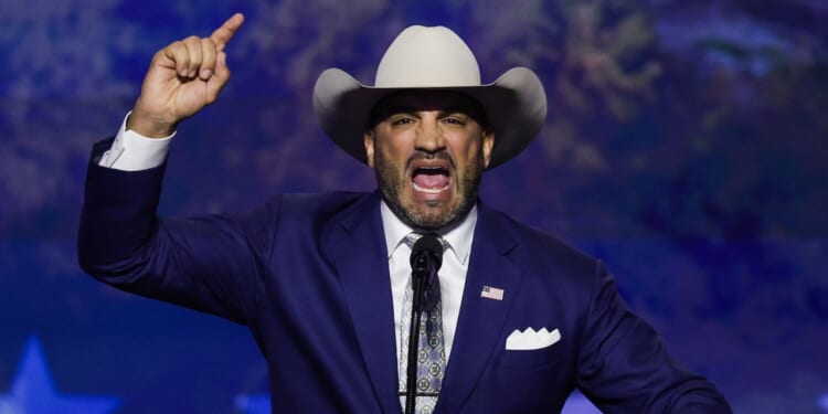 Webb County Judge Tano Tijerina in a blue suit and cowboy hat speaks passionately at a podium, raising his right hand with his index finger pointed upward. An American flag pin is on his lapel, and the background features a blue, starry pattern.
