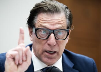 A man in a suit and glasses speaks emphatically, raising his index finger during what appears to be a formal hearing or meeting.