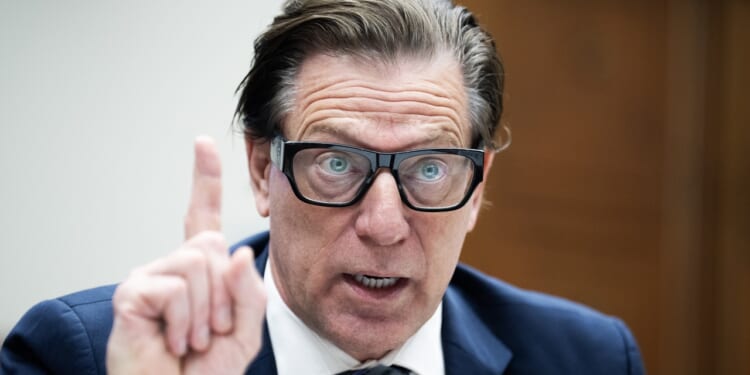 A man in a suit and glasses speaks emphatically, raising his index finger during what appears to be a formal hearing or meeting.
