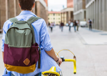A person with a backpack and blue shirt walks their yellow bike down a city street, with buildings and several people in the background.