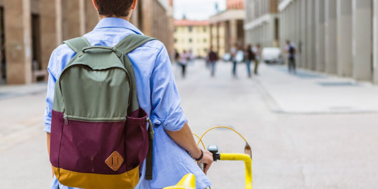 A person with a backpack and blue shirt walks their yellow bike down a city street, with buildings and several people in the background.