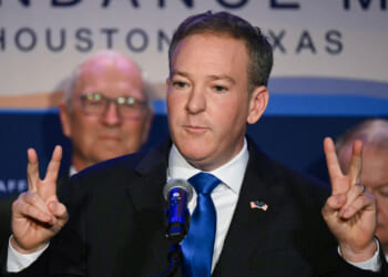 A man in a suit and blue tie stands at a podium, holding up both hands making peace signs, with two men blurred in the background and a banner reading Houston, Texas behind him.