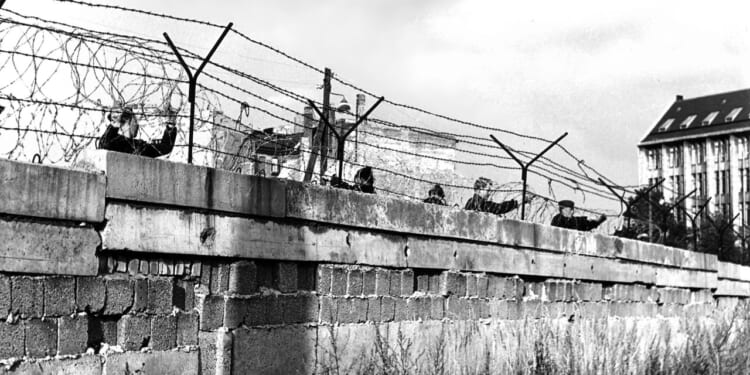 Black-and-white photo of people peering over a concrete wall topped with barbed wire. A building is visible in the background, and grass grows along the base of the wall.