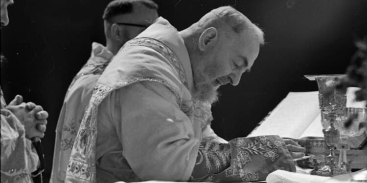 Padre Pio, a bearded priest in ornate vestments leans forward in prayer at an altar, with a chalice and open book in front of him, another clergy member stands nearby.