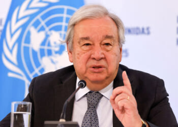 A man in a suit speaks at a podium with a microphone, gesturing with his finger raised. Behind him is a UN logo and a glass of water on the table.