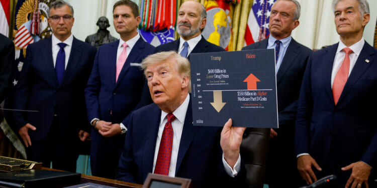 Donald Trump sits at a desk holding a chart comparing drug prices, surrounded by six men in suits, with U.S. flags and military emblems in the background, highlighting his focus on the economy.