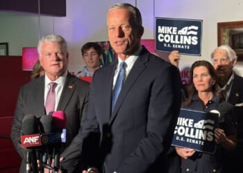 Senator John Thune is pictured in a suit and speaks in front of reporters' microphones. Several people stand behind him holding Mike Collins U.S. Senate signs. The setting is indoors with red lamps.