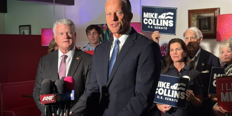 Senator John Thune is pictured in a suit and speaks in front of reporters' microphones. Several people stand behind him holding Mike Collins U.S. Senate signs. The setting is indoors with red lamps.