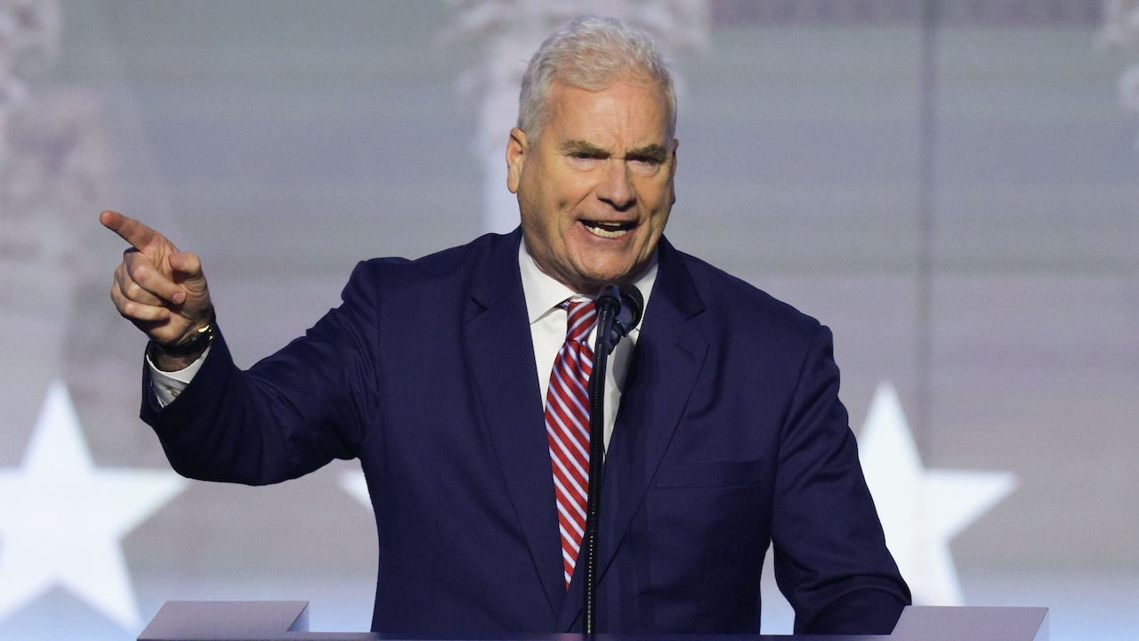 A man in a navy suit and striped tie speaks passionately at a podium, gesturing with his right hand. A blurred background with white star shapes is visible behind him.