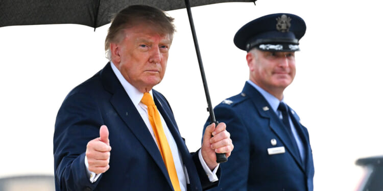 Donald Trump in a suit holds an umbrella and gives a thumbs up, standing next to a uniformed officer outdoors.