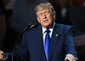 A man in a dark suit and blue tie speaks at a podium with a microphone, gesturing with his hand. An American flag pin is visible on his lapel. The background is dark and blurred.