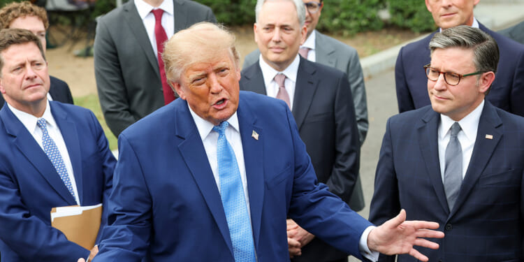 A man in a blue suit speaks animatedly to a group of men in suits, who stand attentively behind him outdoors.
