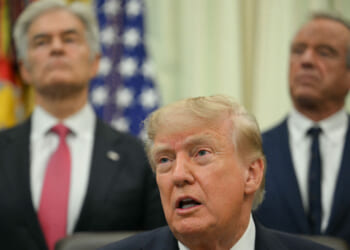 Three men in suits stand indoors; the man in front is speaking, while two others stand behind him. U.S. flags and official emblems are visible in the background.