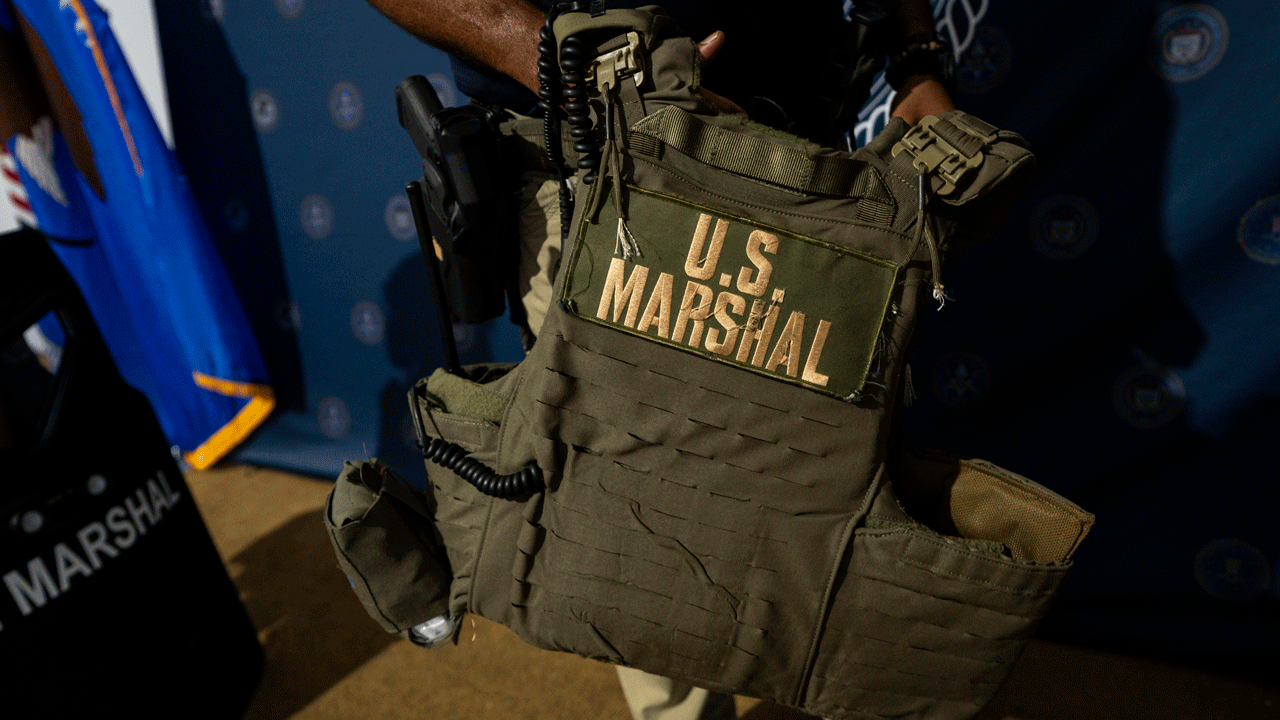 A person holds a tactical vest labeled U.S. Marshal in large yellow letters, with a radio attached. A U.S. Marshals shield and blue flag are visible in the background.