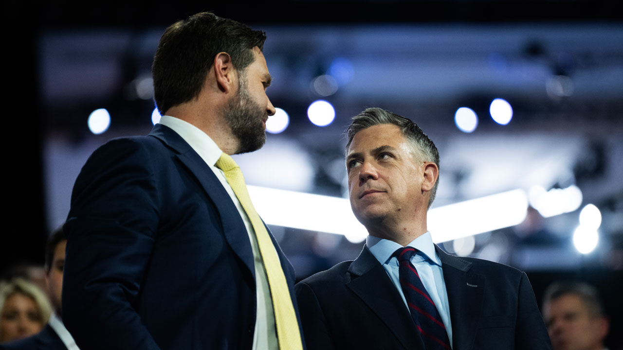 JD Vance and Jim Banks in suits stand close together, facing each other and talking, with a blurred crowd and bright lights in the background.