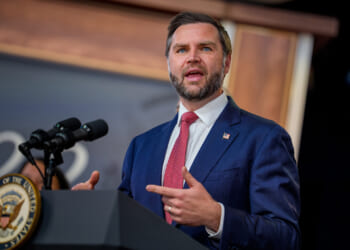 JD Vance in a blue suit and red tie speaks at a podium with microphones; the podium displays the seal of the President of the United States.