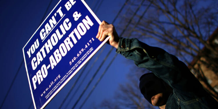 A person holds a large sign reading, You cant be Catholic & pro-abortion, with contact information and a website, against a blue sky and some tree branches.