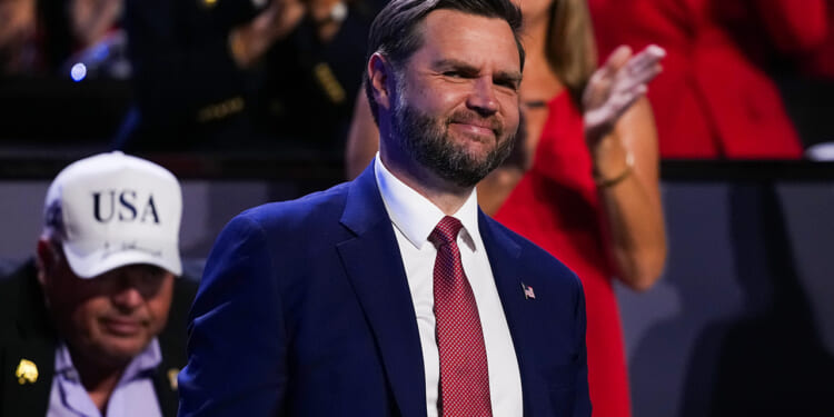 A man in a blue suit and red tie smiles while standing at an event. People are visible in the blurred background, including someone in a white USA hat.