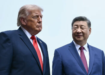 Donald Trump and Xi Jinping in dark suits and red ties stand side by side outdoors. One looks serious, facing forward, while the other smiles gently, looking at his companion. Both have short, neatly styled hair and stand against a pale sky background.