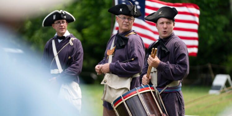 Three men in historical military uniforms stand outdoors with an American flag behind them. One plays a drum, while the others watch. The scene appears to be a reenactment.