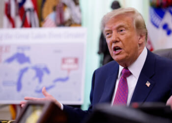 A man in a suit gestures while speaking in an office. Behind him is a blurred map labeled The Great Lakes and some United States flags.