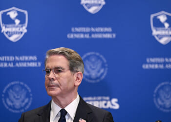 A man in a suit and glasses stands in front of a blue backdrop with United Nations General Assembly and U.S. logos, looking to the side.