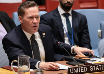 Mike Walz in a suit speaks emphatically at a conference table with a sign reading United States. He has papers and a microphone in front of him, and people are seated nearby.