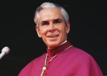 An older man in religious attire with a red robe and gold chain stands at a microphone, smiling slightly, against a dark background.