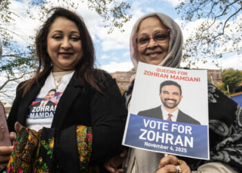 Two women outdoors hold a Vote for Zohran campaign sign and a phone. One wears a shirt with the candidates photo; both are smiling. There are trees and a blue sky in the background.