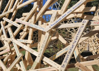 A large, complex sculpture made of interlocking light-colored wooden planks covered in handwritten messages and signatures.