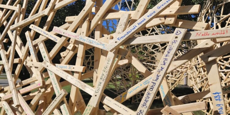 A large, complex sculpture made of interlocking light-colored wooden planks covered in handwritten messages and signatures.