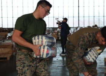 Two service members in camouflage uniforms carry packages of supplies in a large hangar.